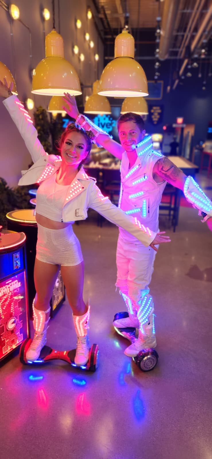 Two performers in white LED jackets and boots riding hoverboards at an arcade venue