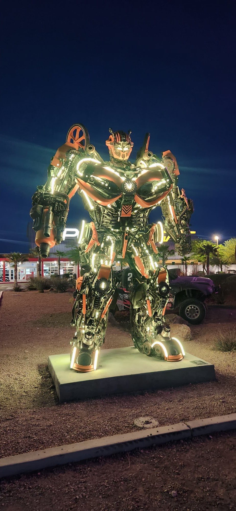 Towering scrap-metal robot sculpture lit with warm LED neon at dusk outside SpeedVegas