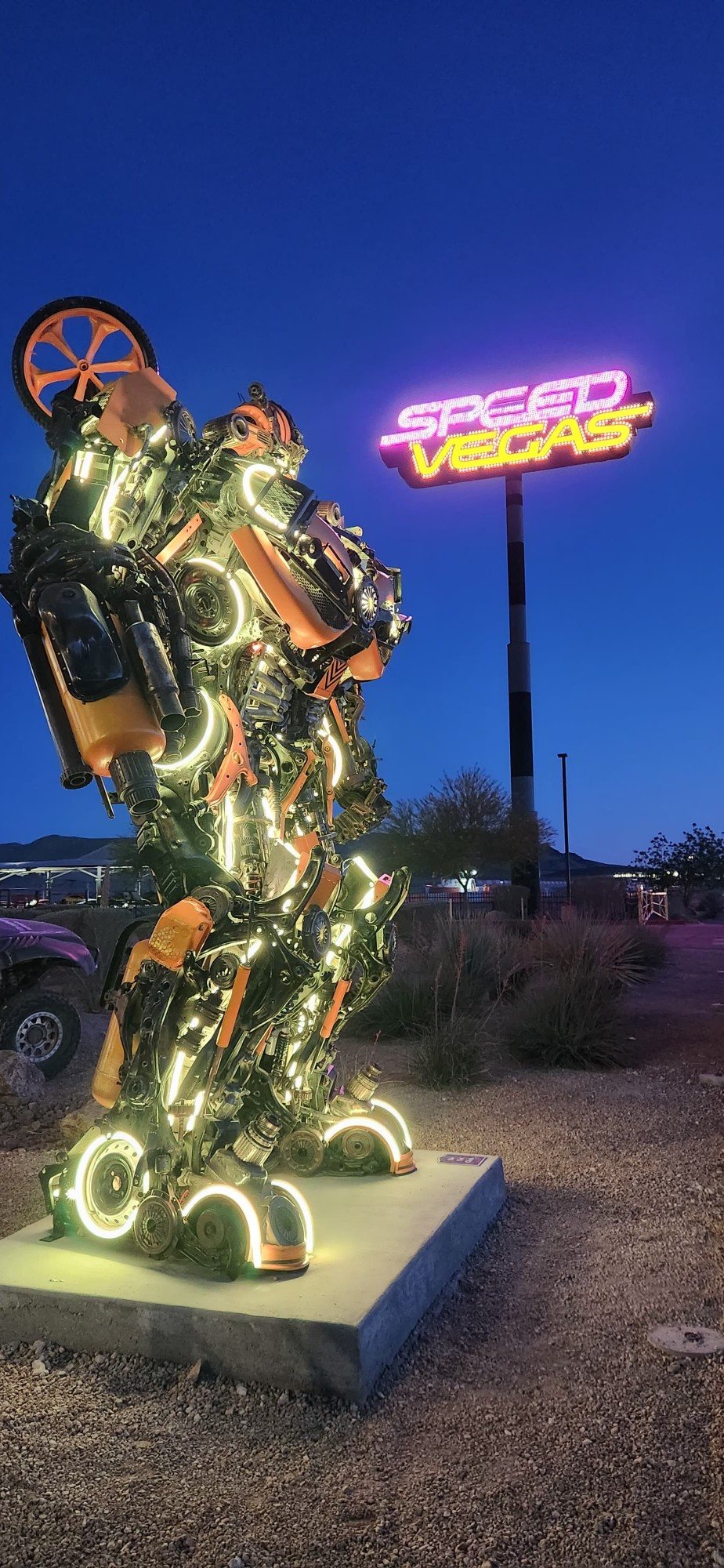 LED-lit scrap-metal robot sculpture in profile with the SpeedVegas sign glowing behind it